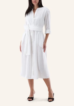 Woman wears white, long-sleeve, button-down midi dress with pleated bodice, tied waist belt, rolled sleeves, and black open-toe heeled sandals.