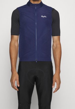 Hombre con chaleco de ciclismo sin mangas azul marino sobre camiseta de manga corta negra y pantalones cortos de ciclismo acolchados negros, de pie frente a un fondo liso.