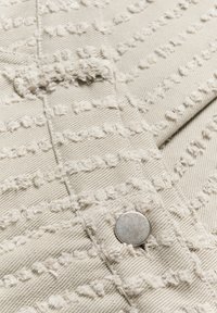 Beige Stoff mit strukturierten Mustern und ausgefransten Details, mit einem silbernen Metallknopf. Das Material scheint mit diagonalen Linien gewebt zu sein.