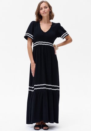 Robe maxi noire à manches courtes, ornée d'une bordure en zigzag blanc à la taille et aux poignets, avec un ourlet évasé. Texture de tissu lisse.