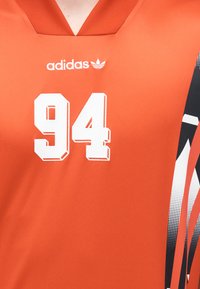 Orange sporttröja med V-ringning, med numret "94" i vitt och svartvita grafiska mönster längs sidan. Adidas-logotyp ovanför.