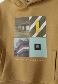 Felpa in tessuto beige con una grafica multi-immagine. Il design include uno skateboard giallo e motivi di architettura urbana con dettagli testuali.