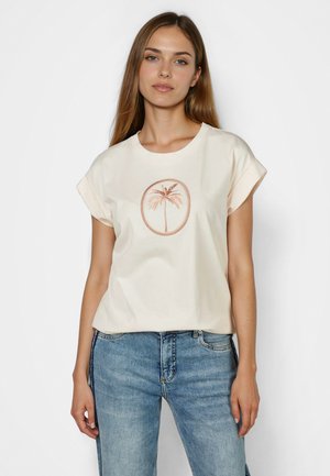 Frau mit langen hellbraunen Haaren, die ein cremefarbenes kurzärmeliges T-Shirt mit braunem Palmenmotiv und blaue Jeans trägt, steht vor einem weißen Hintergrund.