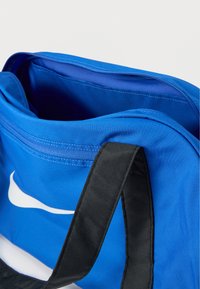 Μπλε αθλητική τσάντα με μαύρα λουριά, που διαθέτει μεγάλο λευκό λογότυπο. Περιλαμβάνει άνοιγμα με φερμουάρ και ομαλή υφή υφάσματος στο εσωτερικό.