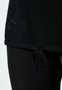 Sort strikbluse med netmønster og sidebindedetalje over tætsiddende sorte bukser på en lys baggrund.