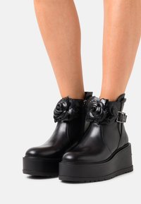 Botas de tobillo de cuero negro con adornos de rosa, cremallera lateral y suela de plataforma gruesa. Detalles texturizados y hebilla ajustable.