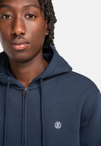 Marineblaue Zip-Hoodie aus Baumwolle, mit einem Frontreißverschluss, einer Kapuze mit Zugband und einem kleinen weißen Logo auf der Brust.