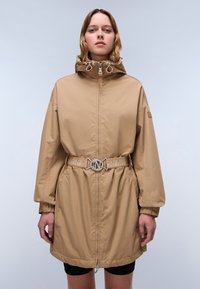 Veste imperméable marron clair avec un col haut, fermeture éclair, taille cintrée et ceinture avec logo. Elle possède des poignets élastiques et des poches latérales.