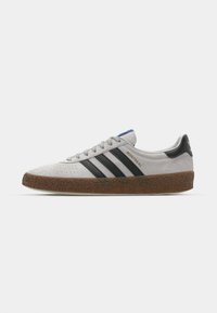 Lys grå ruskinds sneakers med sorte striber og en brun, tekstureret gummisål. Har en blå accent på tungen og et guld "MONTREAL" logo.