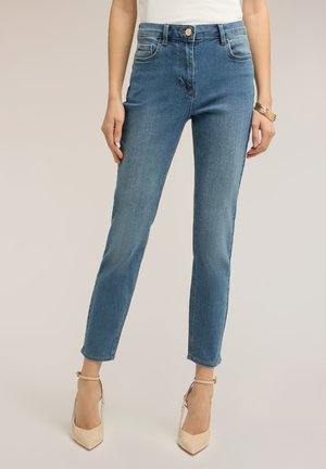 Frau trägt hoch taillierte schmale blaue Jeans, weißes Oberteil, beige Knöchelriemen-Pumps und goldene Armreife am rechten Handgelenk, steht vor einfarbigem Hintergrund.