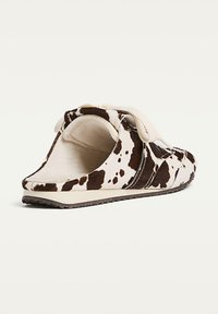 Slip-on shoe with a cow print pattern, made of soft material. Features a beige fabric strap and a textured rubber sole. Back is open.