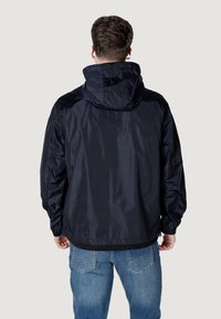 Giacca con cappuccio blu navy realizzata in un tessuto liscio e resistente all'acqua, con polsini elastici e vestibilità comoda. Indossata sopra una maglietta nera e jeans blu.