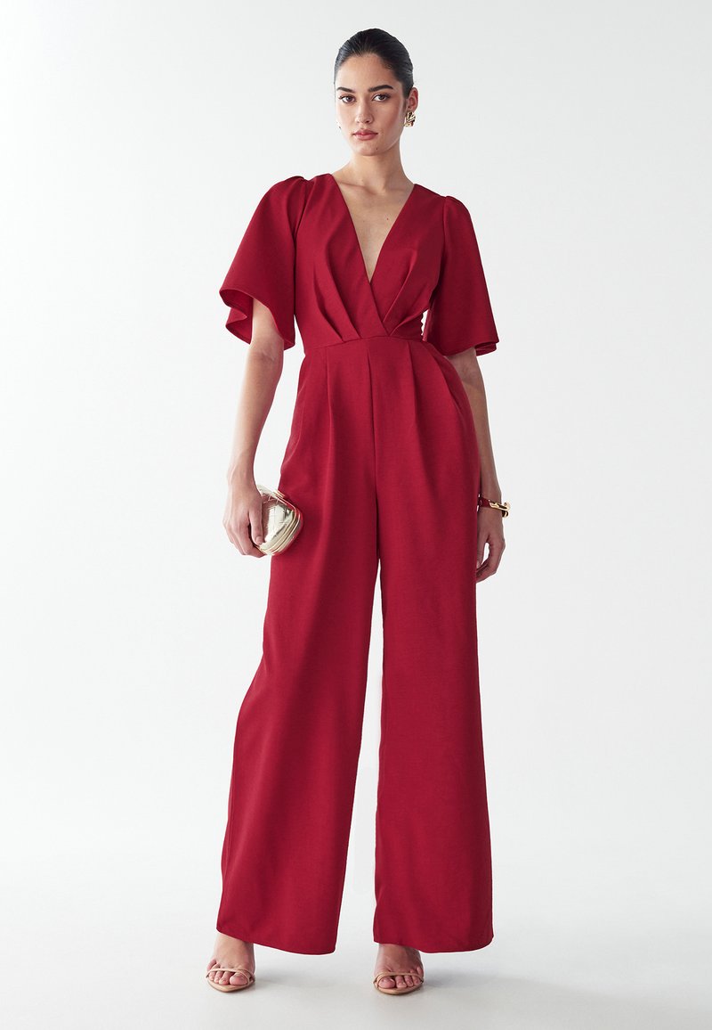 Rode jumpsuit met korte, brede mouwen, diepe V-halslijn, geplooide voorkant en wijde pijpen. Model houdt een kleine metalen handtas vast.