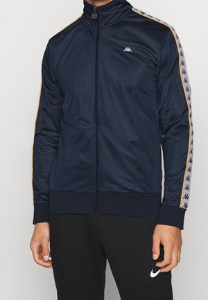 Marine trainingspak jas gemaakt van gladde stof, met een hoge kraag, ritssluiting en gouden accenten langs de mouwen, inclusief logo-detail.