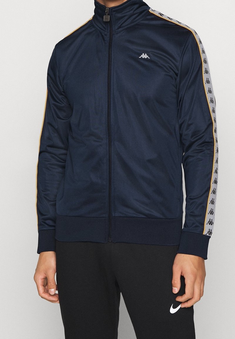 Marinblå träningsjacka i slät tyg, med hög krage, dragkedja och gulddetaljer längs ärmarna samt logotyp.