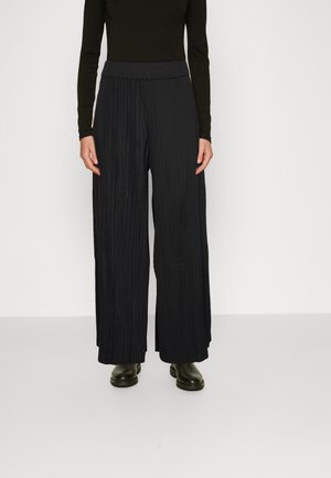 Person wearing black, high-waisted, wide-leg pleated pants paired with a black long-sleeve fitted top and black shoes.