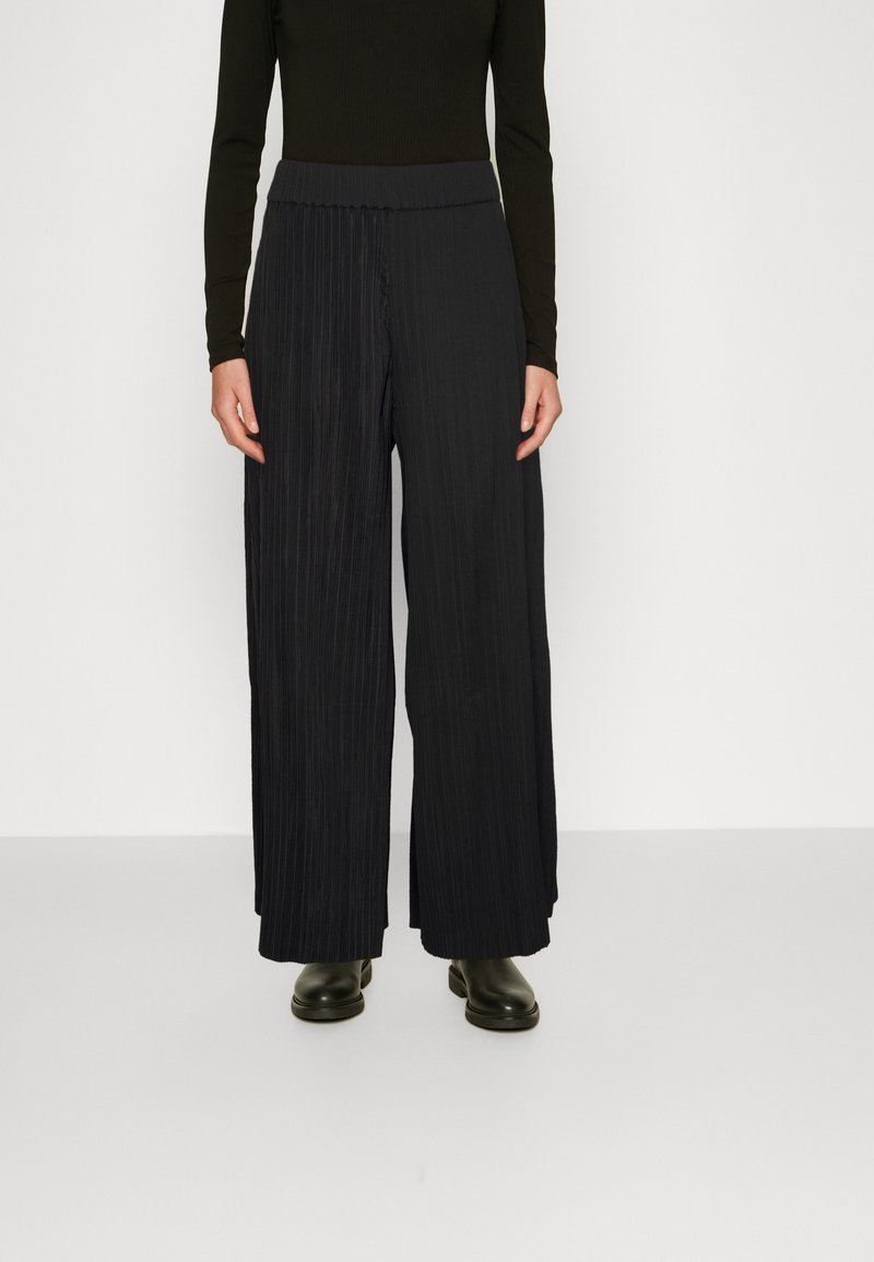 Personne portant un pantalon noir plissé, taille haute et jambes larges, associé à un top noir ajusté à manches longues et des chaussures noires.