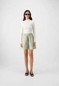 White long-sleeve top, green striped shorts with elastic waist, black heeled mules, and rectangular sunglasses. Simple, modern design.