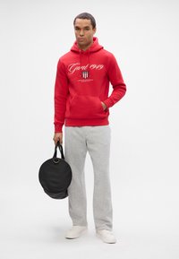 Homme portant un sweat à capuche rouge et un pantalon de jogging gris, tenant un sac rond noir, se tenant devant un fond blanc uni.
