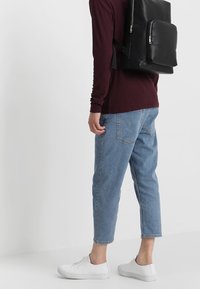 Svart läderryggsäck med framficka med dragkedja, bärs över en vinröd ribbad långärmad topp. Ljusblå avklippta jeans och vita sneakers synliga.