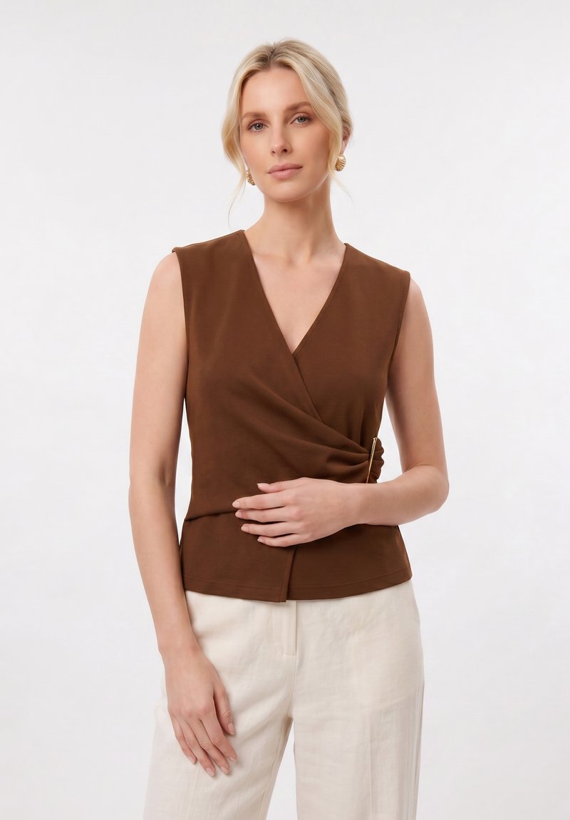 Femme aux cheveux blonds portant des boucles d’oreilles créoles dorées, un haut cache-cœur marron sans manches et un pantalon beige clair, debout devant un fond blanc.