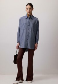 Blå skjorte med knap-op og to brystlommer, parret med brune flarebukser. Outfittet er accessorized med et mønstrede halstørklæde og en mørk håndtaske.