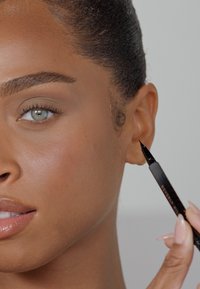 Penna per eyeliner nero tenuta vicino all'orecchio di una persona con pelle liscia e dal tono uniforme, sopracciglia prominenti e occhi azzurri chiari.