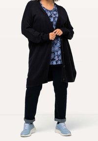 Chaqueta de punto negra con cierre de botones, llevada sobre una blusa de patrón floral azul. Combinada con jeans oscuros y zapatillas azul claro.