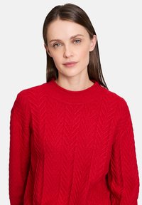 Maglione rosso a maglia con motivo a zigzag, collo rotondo e maniche lunghe. Il tessuto appare texturizzato e caldo, adatto per il clima più fresco.
