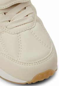 Beige Sneaker mit einem strukturierten, synthetischen Obermaterial, flachen Schnürsenkeln, genähten Akzenten und einer leichten Gummisohle mit abgerundetem Profil für besseren Halt.