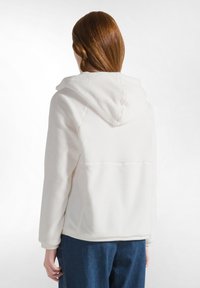 Chaqueta con capucha de forro polar blanca con una textura suave, que presenta un corte de longitud media, un bolsillo canguro y detalles de costura a lo largo de las costuras.
