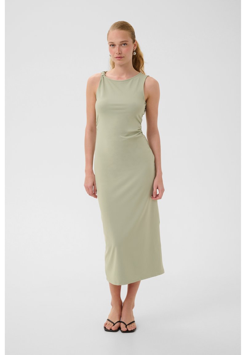 Femme debout portant une robe midi sans manches vert clair avec un détail de nœud à l'épaule et des sandales plates noires sur fond uni.