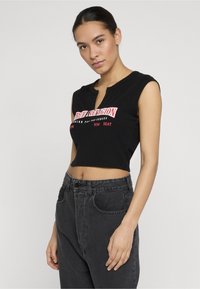 Noor naine, kellel on tagasi seotud tume juuksed, kannab musta varrukateta kõhukest särk punase ja valge tekstiga ning kõrge vöökohaga tumedaid teksaseid.