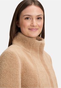 Kobieta z prostymi brązowymi włosami, ubrana w teksturowany beżowy sweter z wysokim kołnierzem, delikatnie się uśmiechająca, na jednolitym jasnym tle.