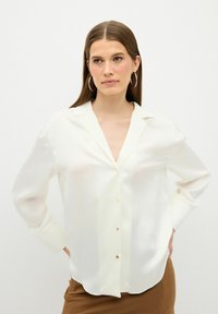 Camicia bianca in seta con colletto, maniche lunghe e chiusura a bottoni, caratterizzata da una vestibilità ampia e una leggera lucentezza. Texture morbida con orlo arrotondato.