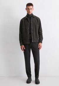 Dark green corduroy jacket, black turtleneck, black textured pants, and black ankle boots. Model stands against a white wall.