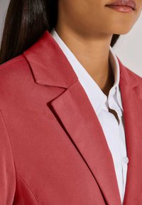 Roter Blazer mit strukturiertem Revers, aus glattem Stoff gefertigt. Kombiniert mit einem weißen Hemd darunter, das ein klares Design zeigt.