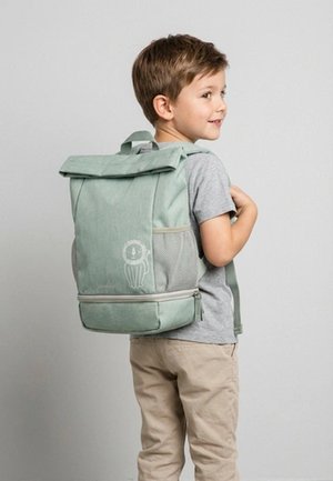 Young boy wearing gray shirt and beige pants carries light green backpack with lion design, standing against plain gray background.