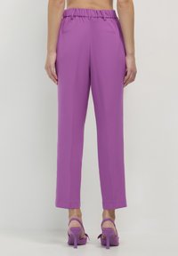 Donna che indossa pantaloni a vita alta viola con tasche posteriori, abbinati a sandali con tacco alto viola, in piedi su uno sfondo semplice.