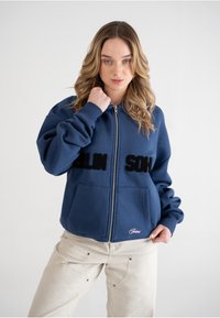 Felpa con zip blu navy, con grandi lettere nere "BERLIN SOHN", tasche frontali e polsini a coste. Tessuto di texture liscia.