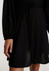 Robe noire avec des manches longues bouffantes et une ceinture à nouer à la taille. Le tissu est léger avec une texture légèrement satinée.