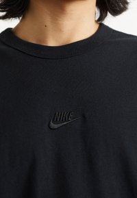 Černé bavlněné tričko s malým černým vyšitým logem Nike na hrudi. Kulatý výstřih a krátké rukávy. Hladká textura.