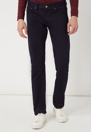 Pantalones slim-fit de color azul marino hechos de algodón, con cinco bolsillos y herrajes de metal cobre, combinados con zapatillas blancas.