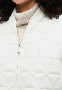 Balta dygsniuota striukė su rombo raštu, su priekiu užtrauktuku ir logotipo akcentu, dėvima ant šviesiai smėlio spalvos megztinio.