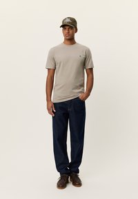 Μπεζ κοντομάνικο μπλουζάκι με διακριτικό λογότυπο, σε συνδυασμό με σκούρο μπλε τζιν παντελόνι με ίσια γραμμή και καφέ δερμάτινα παπούτσια με κορδόνια· ελιάς πράσινο καπέλο.