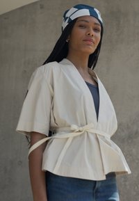 Beige innpakningsbluse med korte ermer, med knyte i midjen og plisserte detaljer; brukt over en mørk topp, sammen med dongeribukser og et mønstrete hodebånd.