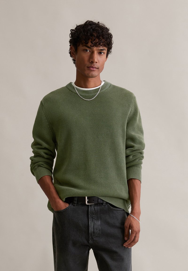 Pulover verde din tricot cu nervuri, mâneci lungi, guler rotund, purtat peste un tricou alb, asortat cu blugi închiși la culoare și o curea neagră. Accesoriu cu lanț argintiu.