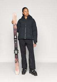Schwarze Steppjacke mit Kapuze, passende schwarze Skihose, hält einen braun-pinken Ski. Schwarze Skistiefel mit strukturierten Akzenten.