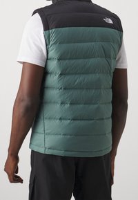 Grüne und schwarze gefütterte Weste mit horizontal gestepptem Muster und einem Logo auf der oberen linken Seite, getragen über einem weißen Hemd.