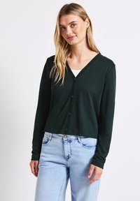 Ciemnozielony krótki kardigan z dekoltem w kształcie litery V, z przodem zapinanym na guziki i długimi rękawami, w połączeniu z jasnoniebieskimi dżinsami. Gładka tekstura.